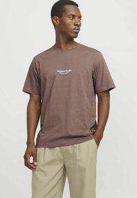 Braunes Baumwoll-T-Shirt mit kurzen Ärmeln, Rundhalsausschnitt und dem Schriftzug "Original Studio" auf der Vorderseite; kombiniert mit hellbeigen Hosen.