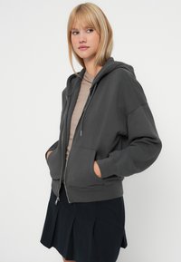 Sivi zip-up dukserica s velikom kapuljačom, prednjim džepovima i rebrastim manžetama, složena preko svijetlo bež majice i kombinirana s crnom suknjom.