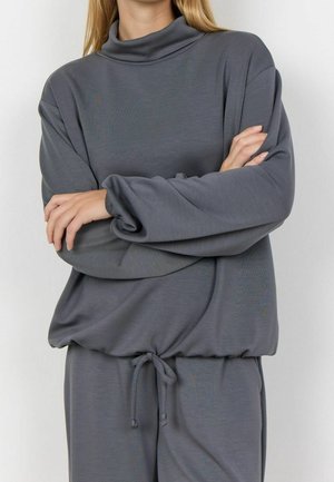 Osoba nosząca luźną szarą bluzkę z długim rękawem i sznurkiem w talii, z założonymi rękami, na jednolitym jasnym tle.