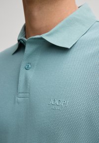 Polo bleu clair en tissu texturé, avec un col à trois boutons et le logo embossé "JOOP! JEANS" sur la poitrine.