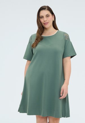 Frau mit langen braunen Haaren, die ein knielanges grünes Kleid mit kurzen Ärmeln und Spitzen-Details auf den Schultern trägt, vor einem schlichten Hintergrund stehend.