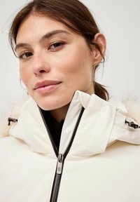 Witte geïsoleerde jas met een hoge kraag, zwarte ritsen en een met bont gevoerde capuchon. Gladde textuur met zijaccenten en minimale branding zichtbaar.