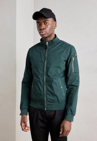 Grønn zip-up jakke med ståkrage, sidelommer og en glidelåslomme p�å armen. Har ribbestrikkede mansjetter og en glatt tekstur.