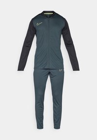 Nike tepláková súprava v tmavom teal a čiernej farbe. Obsahuje plný zips, dlhé rukávy a zúžené nohavice. Ozdobená žltým prešívaním a logo.