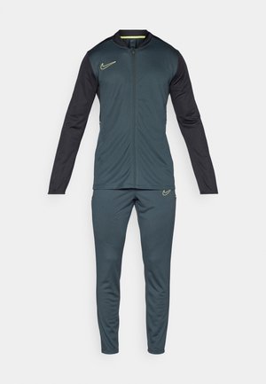 Nike treniruotės kostiumas tamsiai žalias ir juodas. Su pilnu užtrauktuku, ilgomis rankovėmis ir siaurėjančiomis kelnėmis. Papuoštas geltonomis siūlėmis ir logotipais.