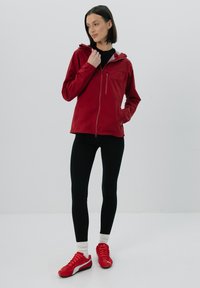 Czerwona lekka kurtka z kapturem, z zamkiem błyskawicznym, zestawiona z czarnymi legginsami oraz czerwonymi butami sportowymi z białymi podeszwami i akcentami.