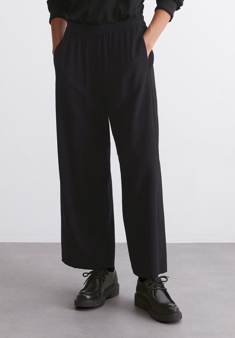 Personne portant un pantalon noir à jambes larges avec ceinture élastique et des chaussures noires à lacets, debout les mains dans les poches sur un sol gris.