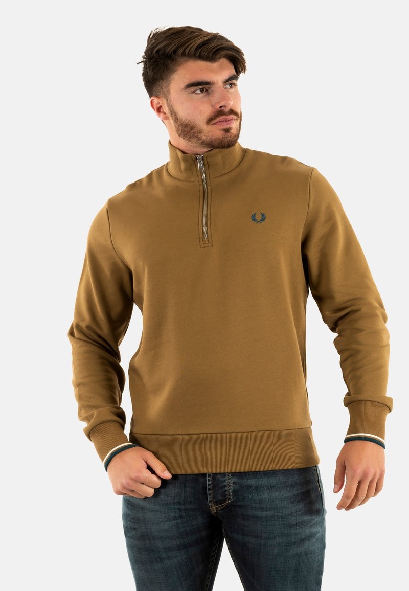 Sudadera marrón con media cremallera, puños y dobladillo de canalé, con un pequeño emblema bordado. Combinada con jeans de mezclilla oscuros. Textura suave.