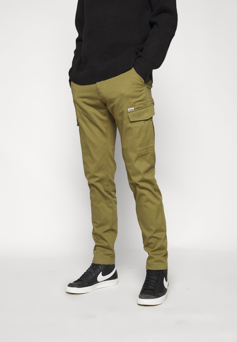 Homme portant un pantalon cargo olive avec poches à rabat et des baskets noires avec semelles blanches et logos Swoosh sur fond blanc.