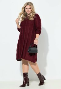 Abito a maglia bordeaux con motivo a trecce, maniche lunghe e vestibilità ampia. Abbinato a stivaletti bordeaux lucidi e una piccola borsa nera.