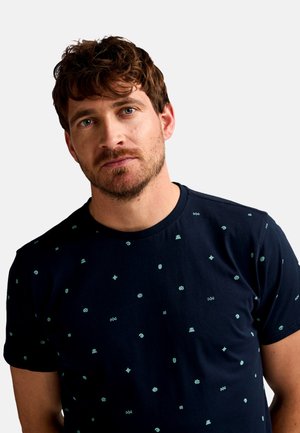 Man met kort bruin haar en baard die een marineblauw T-shirt draagt met kleine lichtblauwe abstracte patronen op een effen achtergrond.