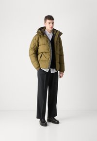 Oliwkowa kurtka puchowa z kapturem, zapinana na zamek, z dwoma bocznymi kieszeniami; noszona na czarnym swetrze i białej koszuli, zestawiona z czarnymi spodniami.