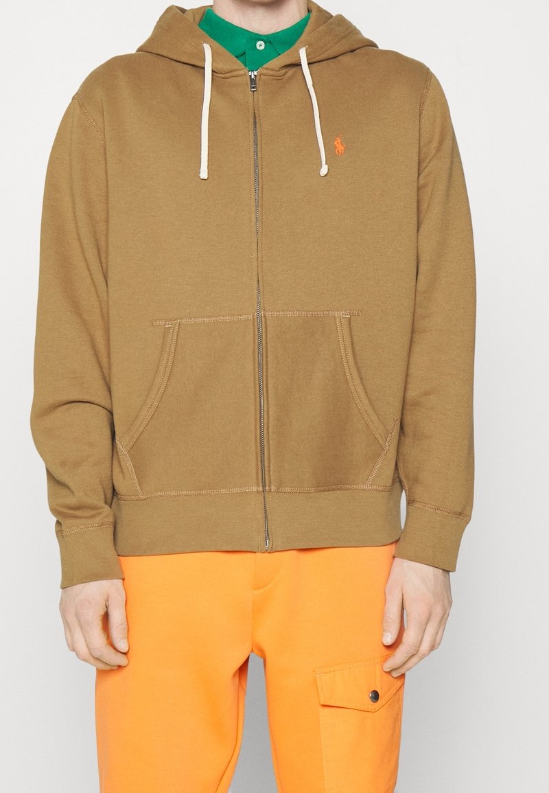 Homme portant un sweat à capuche marron zippé avec poches avant, chemise verte en dessous, et pantalon orange vif avec poche latérale.