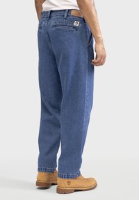 Jeans en denim bleu avec une coupe droite, dotés d'une patch en cuir sur la taille, de poches latérales et de détails de couture. Assortis avec des bottes beiges.