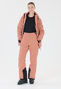 Roze geïsoleerde skijas met een capuchon, zwarte accenten en verstelbare banden. Bijpassende roze skibroek met een zwart paneel op het onderbeen. Zwarte skibotten.