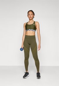 Olívazöld sportmelltartó fekete "RUNDER" pánttal, hozzáillő magas derekú leggings-szel, sárga részletekkel. Kék súlyzót tart.