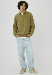 Un jeune homme se tient les mains dans les poches, portant un pull zippé olive, un jean bleu clair et des chaussures beiges à lacets, devant un fond uni.