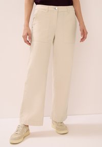 Béžové široké cargo kalhoty s předními kapsami, zapínáním na knoflík a poutky na pásek, vyrobené z lehkého materiálu. S kombinací béžových tenisek.