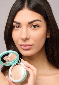 Kompaktpuder in einem türkisfarbenen Etui mit einem Spiegel; wird von einer Person mit glatter Haut und neutralem Make-up gehalten, die eine lichtreflektierende Oberfläche zeigt.
