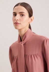 Blusa rosa blush con colletto a fiocco, parte anteriore plissettata e maniche a sbuffo in un tessuto liscio e leggero. Nessun motivo o hardware visibile.