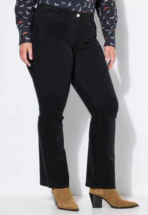 Persona con pantalones negros ajustados, una camisa oscura con pequeños estampados de perros y botas de tacón beige hasta el tobillo, de pie contra una pared blanca.