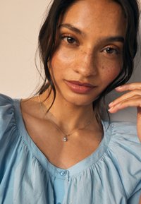 Hellblaue Bluse mit Puffärmeln und Knopfdetail, silberne Halskette mit rundem Anhänger, glatte Haut mit Sommersprossen.