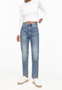 Celana jeans biru muda dengan pinggang tinggi, potongan lurus, lima kantong, dan tekstur yang pudar, dipadukan dengan atasan putih lengan panjang cropped dan sepatu sneakers putih.