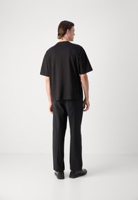 T-shirt nera in cotone con vestibilità rilassata e maniche corte, abbinata a pantaloni scuri e scarpe nere, vista da dietro.