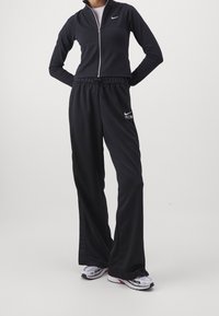 Svart träningsset som innehåller en zip-up jacka med långa ärmar och lösa, dragsko-byxor. Båda delarna har en vit Nike-logotyp som accent.