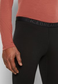 Black leggings with a smooth texture and a black waistband featuring the word "ICEBREAKER" in gray. Worn with a long-sleeve, rust-colored top.