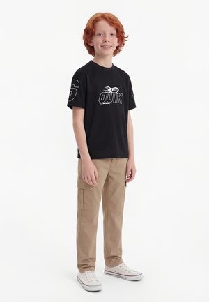 Rothaariger Junge steht, trägt ein schwarzes T-Shirt mit Grafik, beige Cargohosen und weiße Turnschuhe, lächelt in die Kamera vor weißem Hintergrund.