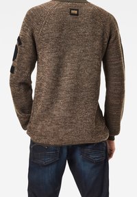 Mann mit einem braunen Strickpullover, auf dem der schwarze Aufdruck "3" am Ärmel und dunkblauen Jeans, von hinten gezeigt.