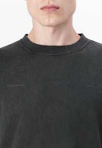 Άνδρας που φοράει σκουρόχρωμη μπλούζα με στρογγυλή λαιμόκοψη, με ανάγλυφα λογότυπα "arkk copenhagen", ένα στυλιζαρισμένο έμβλημα και τη φράση "est 2015" στο στήθος.