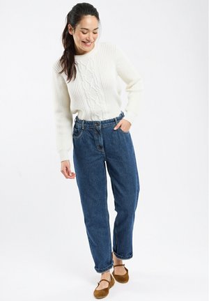 Un pull en tricot blanc associé à un jean en denim bleu taille haute et ample. Des chaussures marron complètent cette tenue décontractée.