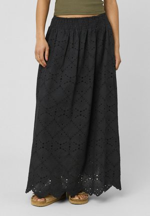 Femme portant une jupe en dentelle œillet noire longue jusqu'aux chevilles avec un ourlet festonné et des sandales compensées couleur beige, vue de la taille vers le bas.