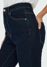 Hoge taille donkerblauwe spijkerbroek met contrasterende gele stiksels, vijf zakken en een gladde denimtextuur. Voorzien van een sluiting met een knoop aan de voorkant.