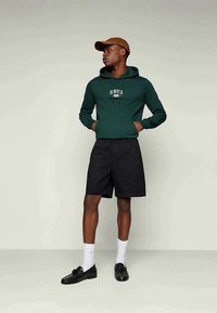 Green hooded sweatshirt with white graphic text, black shorts, white socks, and black loafers. Brown cap completes the casual outfit.