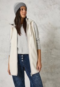 Crème fleece vest met rits, grijze langemouwen shirt eronder, denim jeans met knoopaccenten en een grijs gebreid beanie.