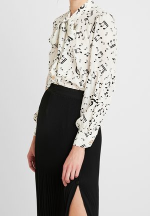 Blouse blanche avec des notes de musique noires et un col nœud, assortie à une jupe plissée noire avec une fente devant et un détail de bouton.