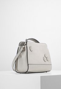 Vaaleanharmaa nahkakäsilaukku, jossa on rakenteellinen muoto, kukkaiset koristeet ja kontrastoiva musta reuna. Mukana säädettävä olkahihna.