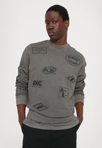 Šedá mikina s různými embosovanými nášivkami s logem "Armani Exchange", kulatý výstřih, proužkované manžety a uvolněný střih.