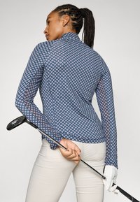 Langärmliges, tailliertes Golfhemd mit einem navyblauen Hintergrund und einem Rautenmuster in Pink und Weiß. Kombiniert mit hellen Hosen.