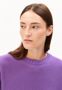 Eine Frau mit hellen Sommersprossen und braunem Haar trägt einen lila Pullover und blickt nach links vor einem weißen Hintergrund.