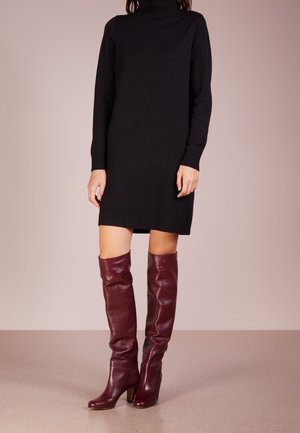Femme portant une robe noire à manches longues avec col roulé et des bottes en cuir rouge foncé montant jusqu'aux genoux, debout sur un sol neutre.