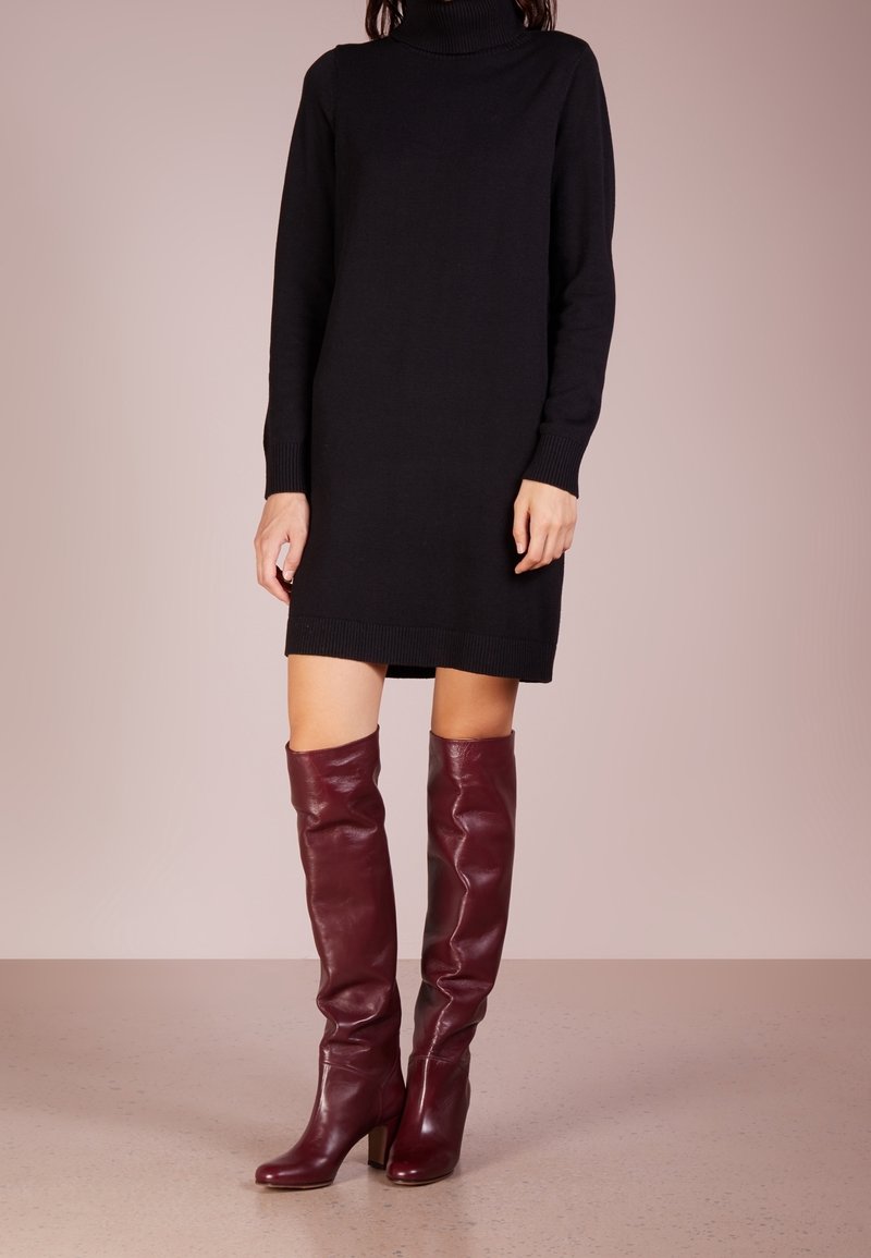 Femme portant une robe noire à manches longues avec col roulé et des bottes en cuir rouge foncé montant jusqu'aux genoux, debout sur un sol neutre.