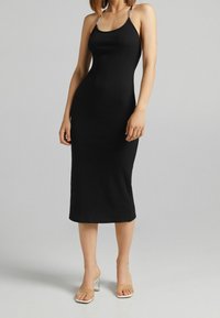 Robe midi noire côtelée avec fines bretelles ornées. Silhouette ajustée, texture lisse, et ourlet arrivant au milieu du mollet. Sandales à talons transparents.