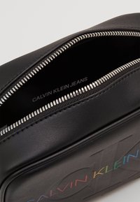 Černá kožená taštička se stříbrným zipem, uvnitř s logem "CALVIN KLEIN JEANS". Hladký povrch a minimalistický design s jemným barevným detailem.