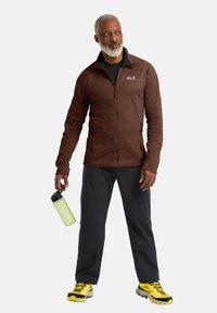 Braune Zip-Jacke, schwarzes Basisteil, schwarze Hose und gelbe Wanderstiefel. Hält eine grüne Flasche. Leicht, athletischer Schnitt, strukturierter Stoff.
