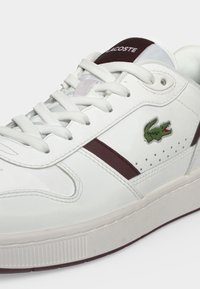 Λευκό sneaker Lacoste με λευκά κορδόνια, μπορντό λεπτομέρειες και το πράσινο λογότυπο κροκόδειλου στην πλευρική επιφάνεια.