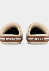 Ζευγάρι μπεζ Crocs χωρίς κορδόνια με καφέ και λευκές ριγέ λωρίδες στη φτέρνα, εμφανιζόμενα από πίσω σε λευκό φόντο.